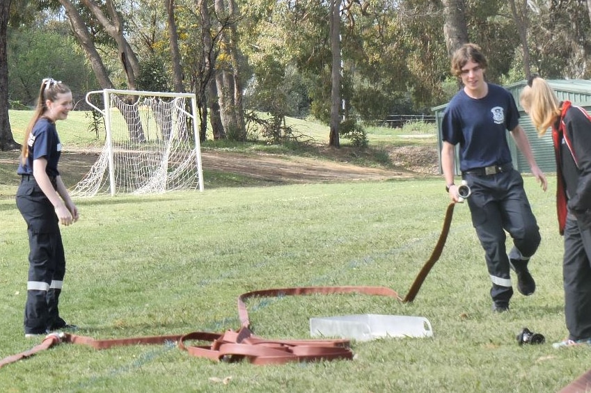 Three students uncoil a red firefighting hose on a school oval with a soccer goal behind them.