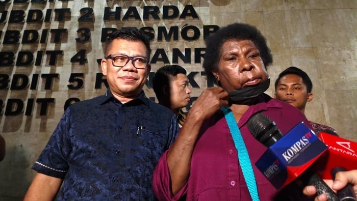 A woman with short curly hair wearing a magenta shirt is accompanied by a man in a blue shirt and glasses.