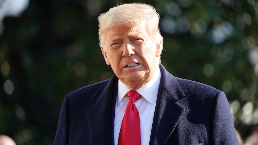 A man in a blue suit jacked and red tie looks at the camera as he speaks to media.