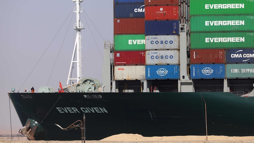 sand in foreground, side view of ship with stacks of coloured containers