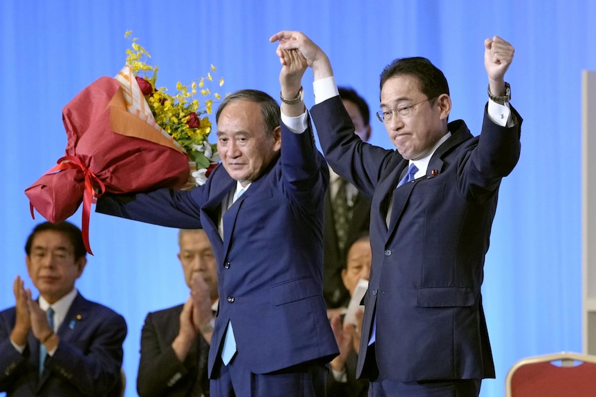 Fumio Kishida and Yoshihide Suga stand on a stage with their hands in the air.