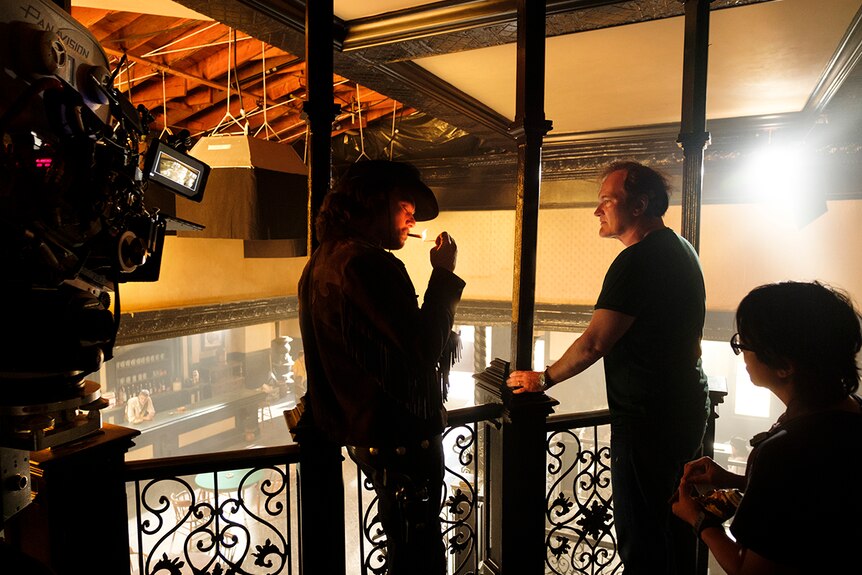 A cinema camera points at two standing men, one lighting a cigarette, on the 2nd floor of a spaghetti western film set saloon.