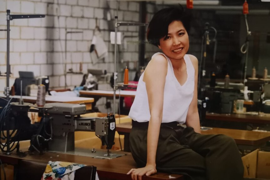 A young woman in a white sleeveless top poses sitting on the sewing machine table inside a garment factory.