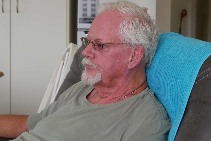An elderly man with grey hair and glasses sits on a lounge chair in his home and looks away from the camera.