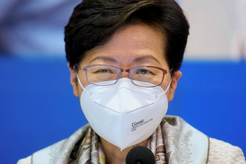 A woman with short dark hair wearing a white P2 facemask and red glasses looks straight ahead.