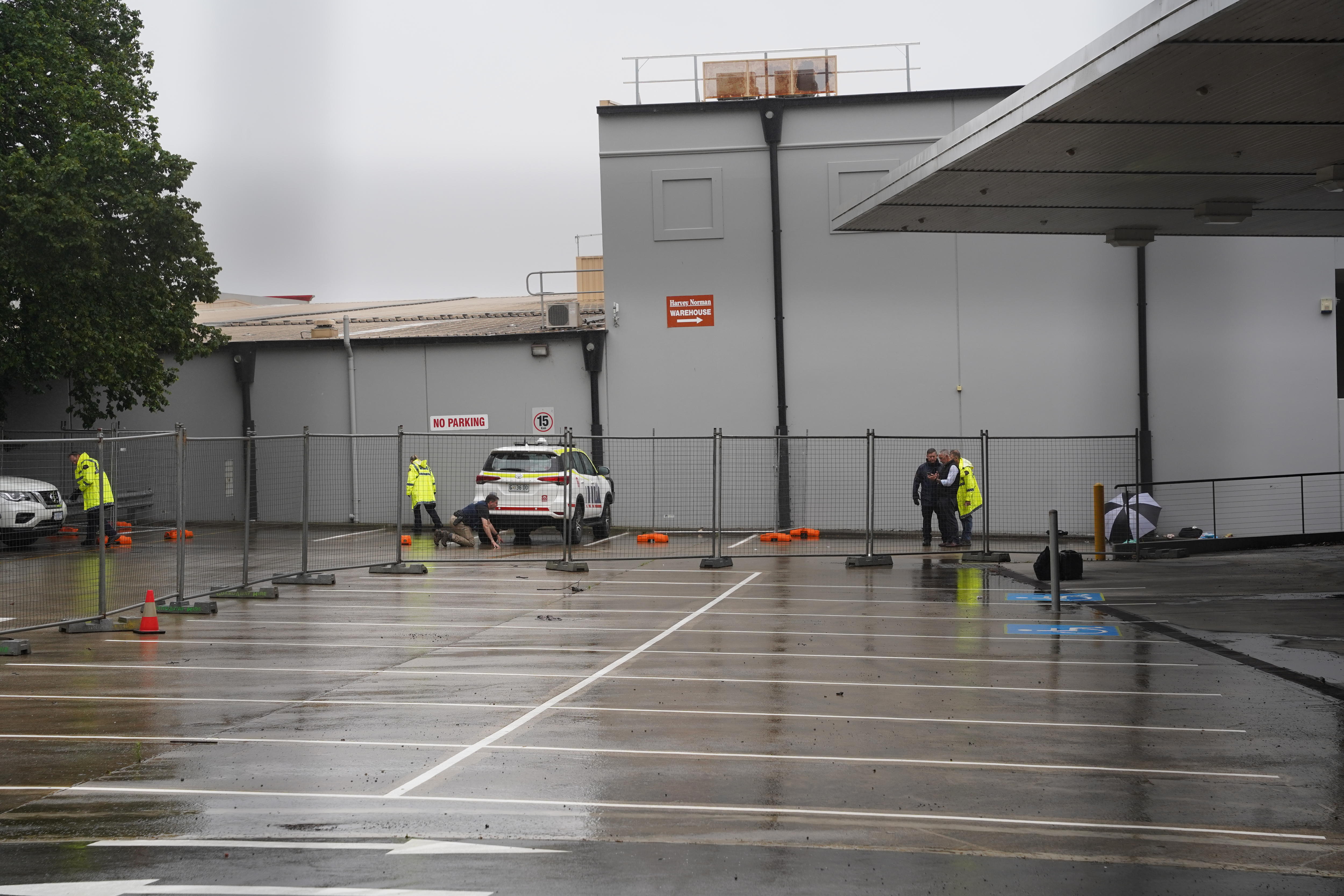 Four police officers wearing hi-vis rain jackets move about a grey, rainy car park with a chain-link fence behind them