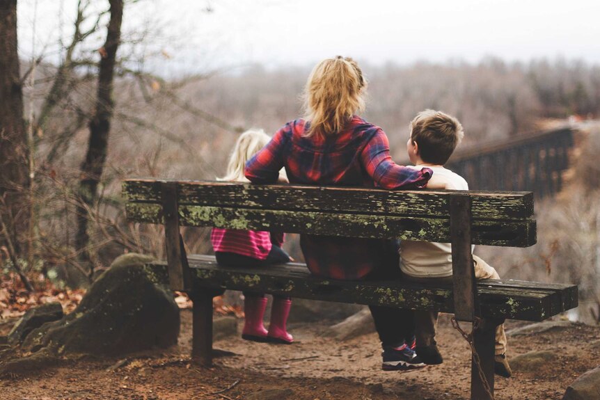 A women with red hair sits on a bench with a child on both sides. She has arms around the two kids