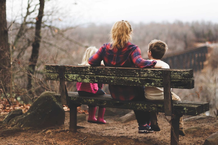 A blonde woman sits in the middle of the bench with two children either side of her. The bench is in a bleak woodland setting