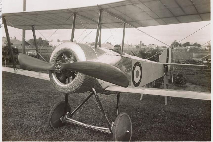 A man sits in the cockpit of an old-fashioned biplane in a suburban garden.