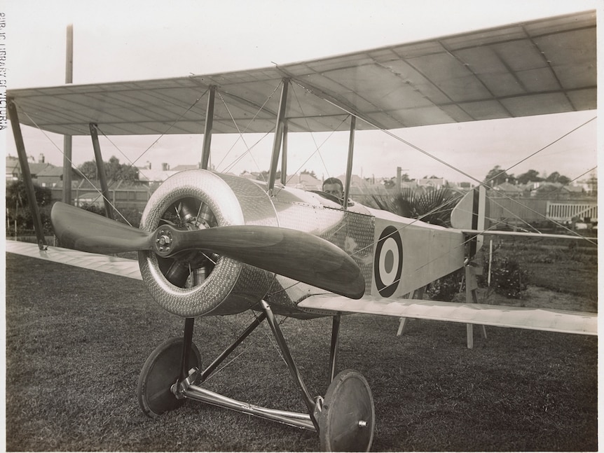 A man sits in the cockpit of an old-fashioned biplane in a suburban garden.