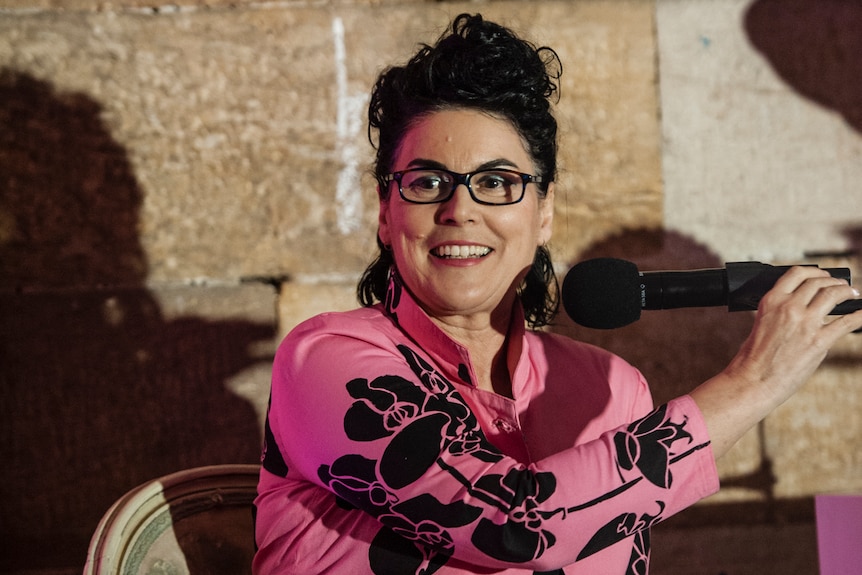 A woman with dark hair in a bun, wearing glasses and a hot pink jacket on smiles broadly as she reaches for a microphone