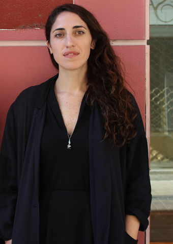 41-year-old woman with long brown curly hair wearing black stands against pink tiled wall.