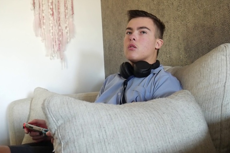 A boy with headphones around his neck sits on a couch in a school uniform.