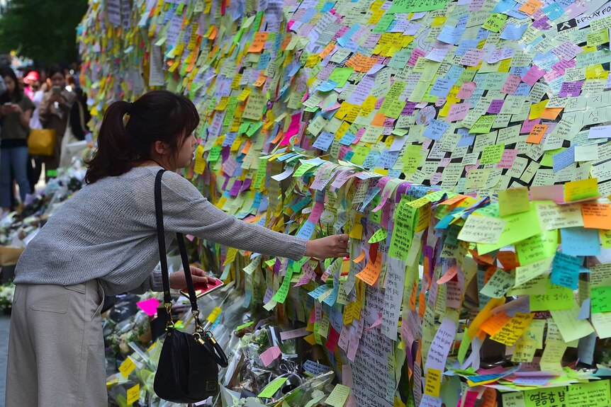 A woman leaves overs to put a post-it note on a wall of multicoloured post-it notes.
