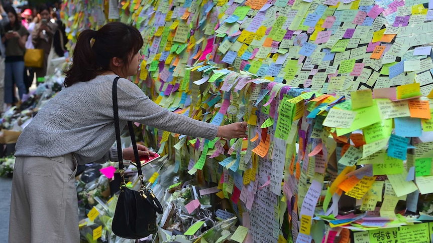 A woman leaves overs to put a post-it note on a wall of multicoloured post-it notes.