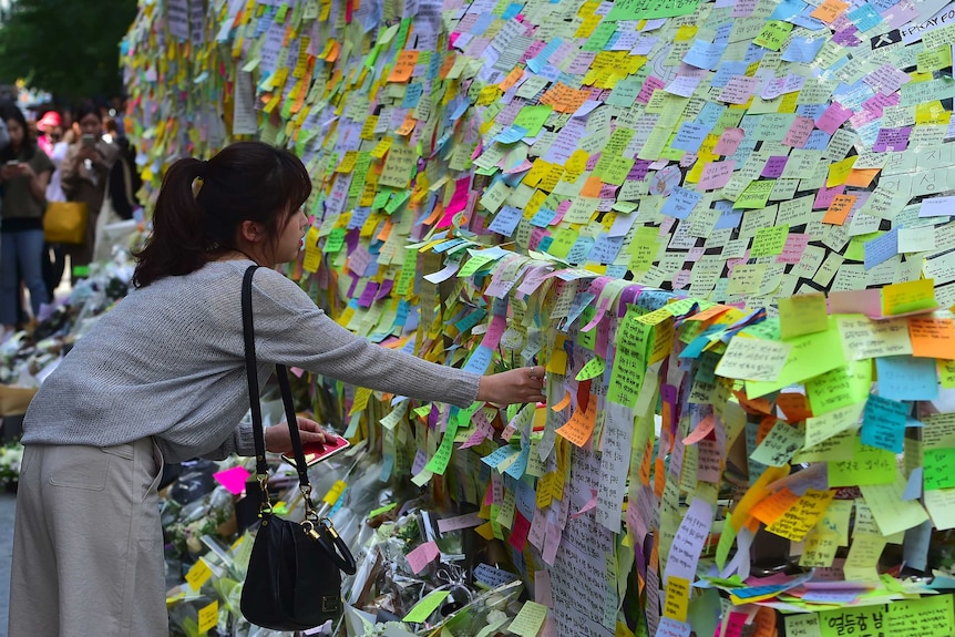 A woman leaves overs to put a post-it note on a wall of multicoloured post-it notes.