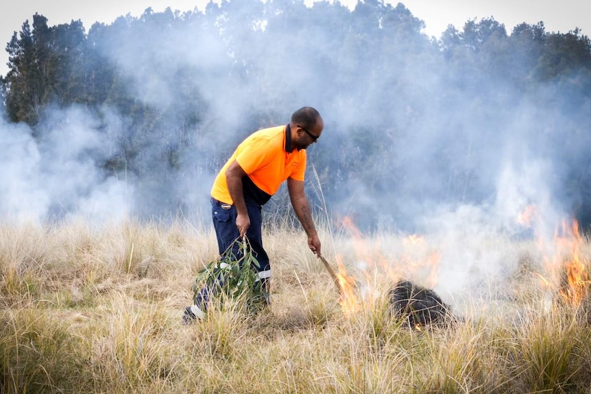 Man surrounded by gentle smoke holding a clump of grass on fire against the ground to light it.