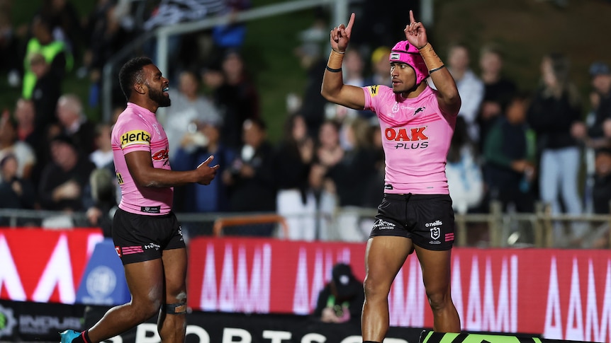 A Penrith NRL player wearing a pink jersey and headgear points his fingers to the sky in celebration near a teammate.