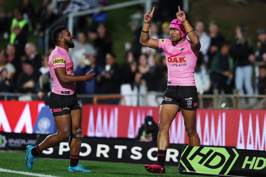 A Penrith NRL player wearing a pink jersey and headgear points his fingers to the sky in celebration near a teammate.