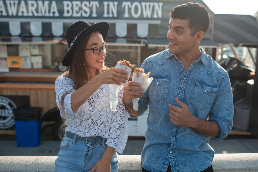 A woman and a man stand next to each other outside, smiling and cheersing their shwarma wraps