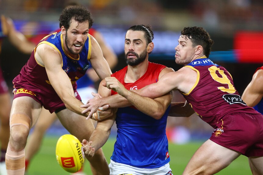 Brodie Grundy handballs from his knees while two Lions players try to tackle him