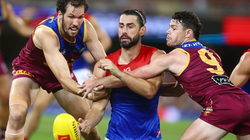 Brodie Grundy handballs from his knees while two Lions players try to tackle him