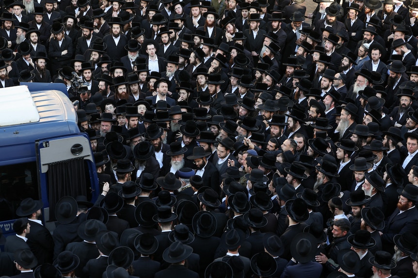 A car is seen in the left side of a large crowd of Jewish men dressed in black