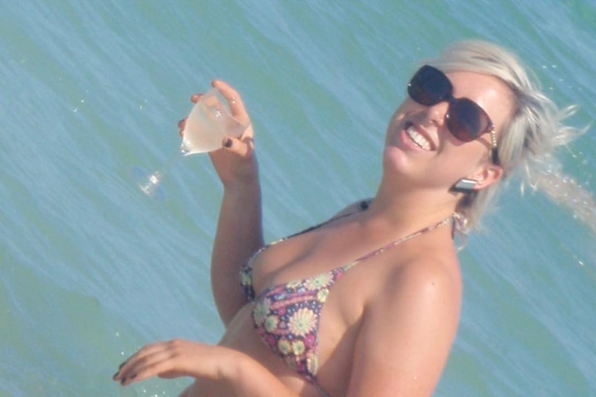 A woman with blonde hair and sunglasses in the ocean with a glass of wine