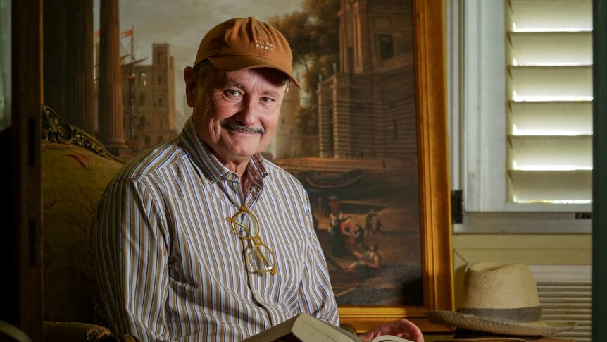U3A member Phillip Beard sits down in his decadent study, holding a book and smiling in front of an ornate painting.