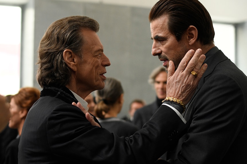 In busy room, an older man in dark suit puts hand with gold jewellery on cheek of taller man with hand on shoulder of older man.