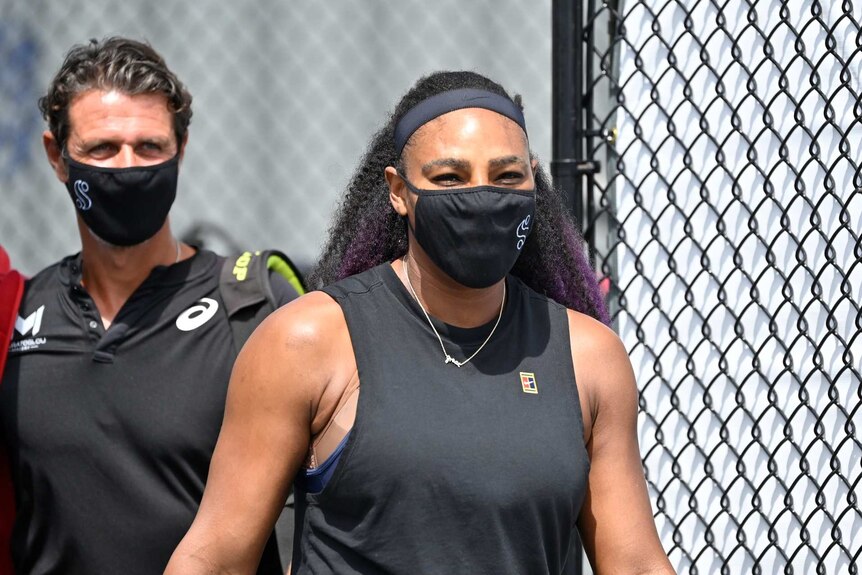 Serena Williams wears a black face mask with a white S on the side as she walks on to a tennis court