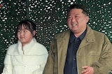Kim Jong Un wearing a black shirt and jacket stands next to his daughter in a white coat.