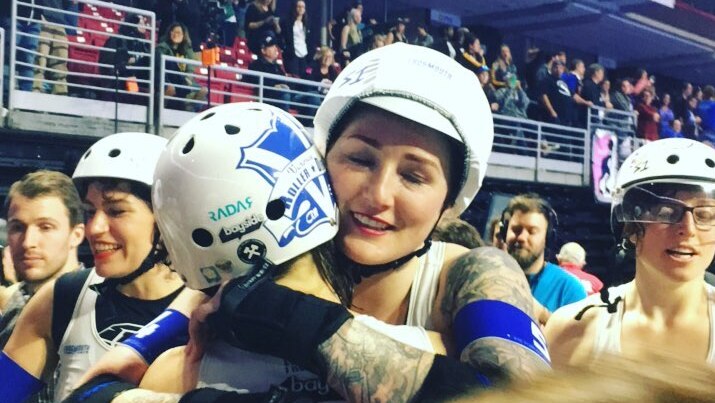 A woman wearing white and blue team colours, helmet and protective padding hugs a team mate as a crowd watches from the stands.