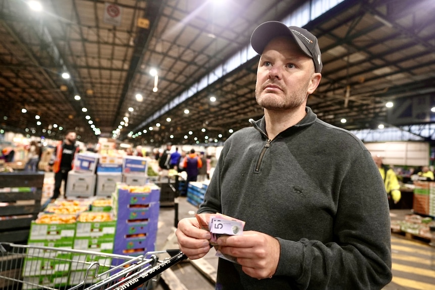 a man in a quarter zip and cap holds a five dollar note looking while shopping for produce