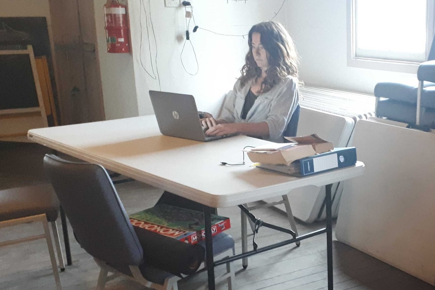 a woman at latop computer at table in a hall, surrounded by stacked chairs