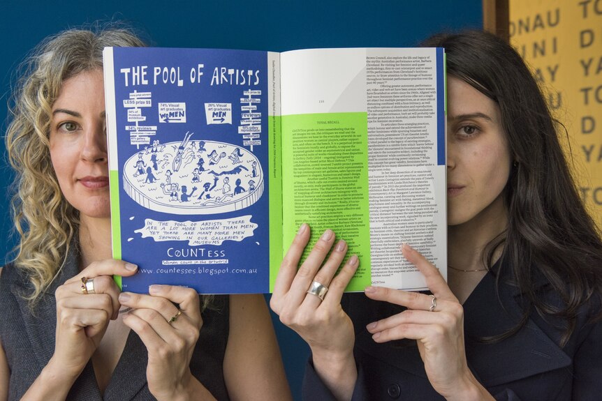 Two women peak out from behind a book they are holding open, which reads "the pool of artists".