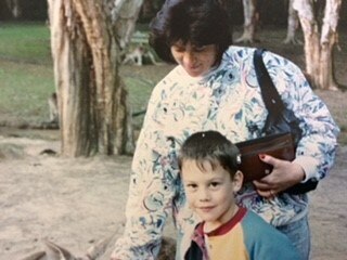A young boy and a woman in a swirling blue and white jacket in a park. Paperbark trees in the background. 