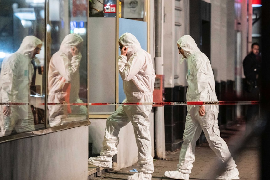 Two men in white protective suits enter a building surrounded by police tape.