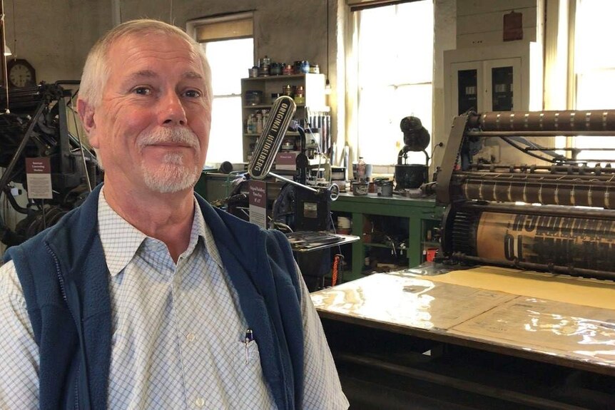 Middle-aged man stands in front of old printers in printing workshop in checked shirt and blue vest