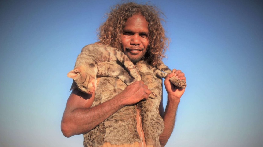An Aboriginal man has a feral cat slung over his shoulders, wearing cat skin vest