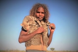 An Aboriginal man has a feral cat slung over his shoulders, wearing cat skin vest