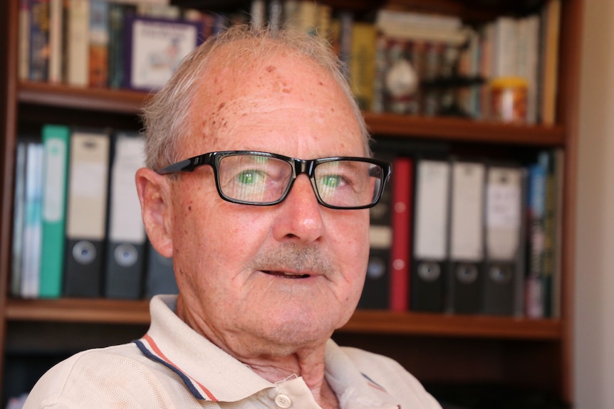 Head shot of an older man wearing glasses staring at camera with a bookshelf behind him.