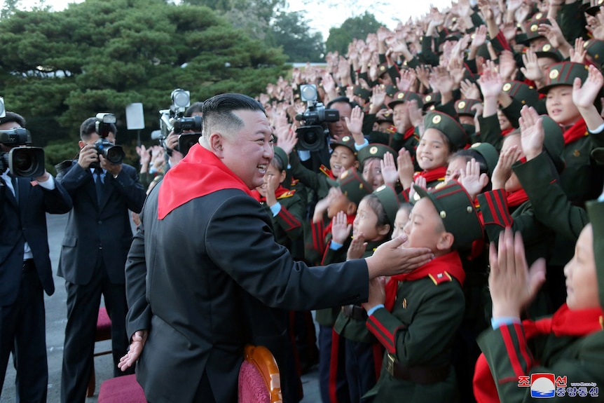 Kim Jong Un stands in front of a crowd of children smiling and patting one child on the face.