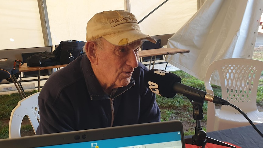 A man in a beige cap sits at a desk speaking into an ABC microphone 