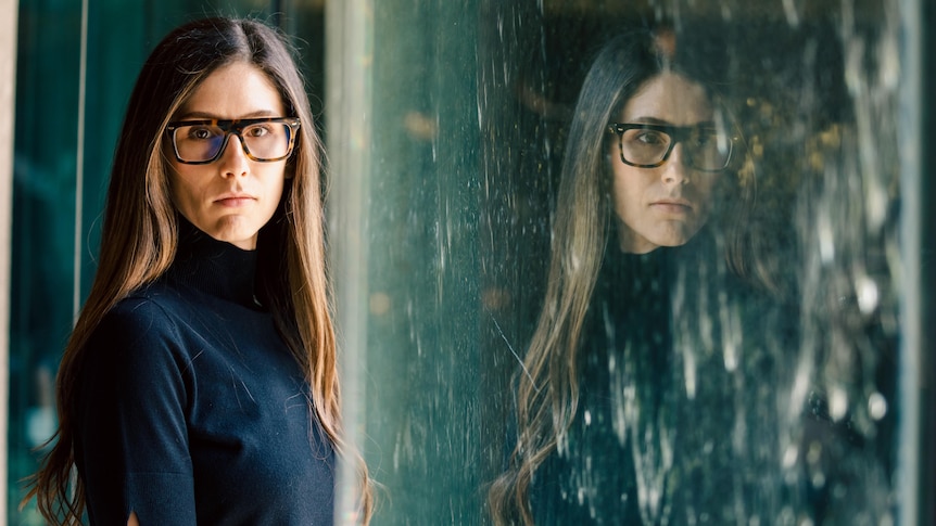 A woman with long hair, a black top and dark framed spectacles poses for a photo in front of a reflective wall of water