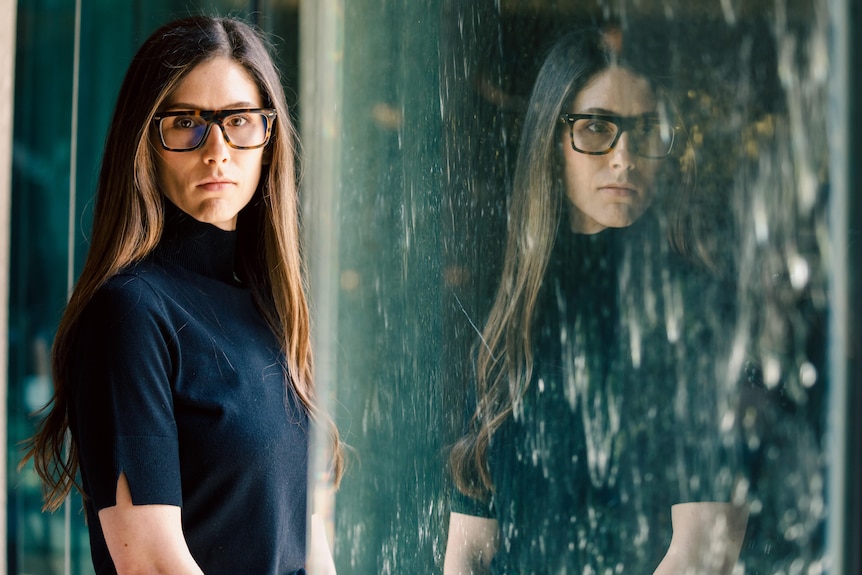A woman with long hair, a black top and dark framed spectacles poses for a photo in front of a reflective wall of water