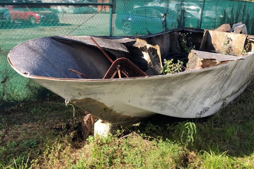 Small boat with weeds and junk in it sits in front of a green mesh fence on grass.