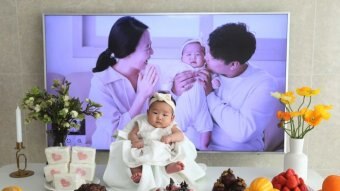 A baby girl sits on a table in white with fruits and a strawberry cake around her. A picture of her parents in the background.