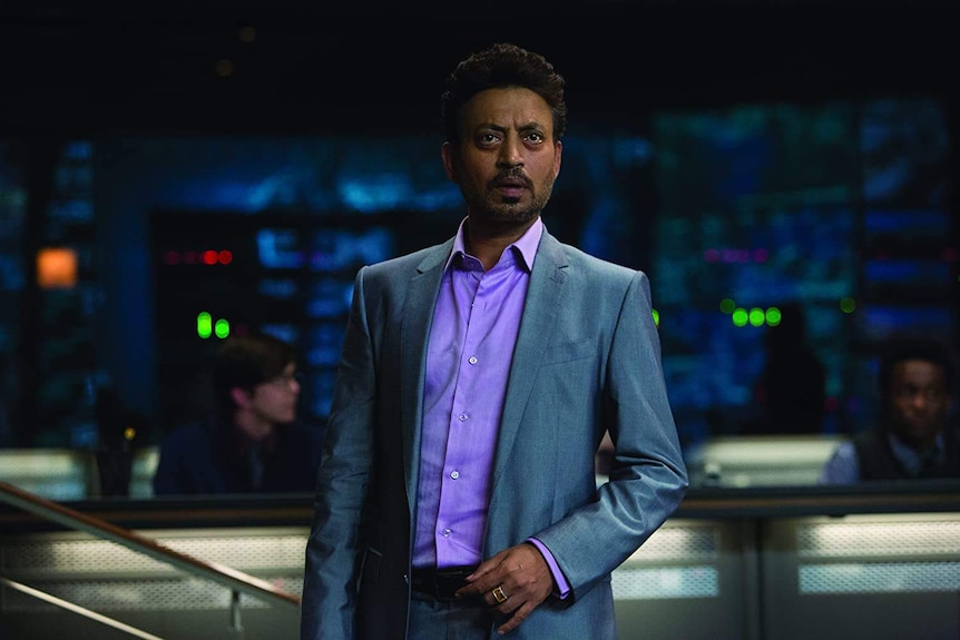 A man stands staring in front of him wearing a grey suit and purple shirt.