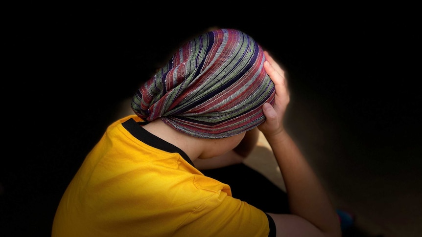 Woman in yellow shirt sits with head, tied in headscarf, in her hands, with black background.